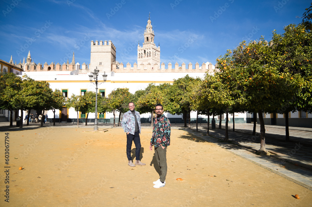 Naklejka premium Young couple of gay men, walking in seville between orange trees, in the background you can see the gothic style cathedral and blue sky. The couple are married. Concept of gay rights