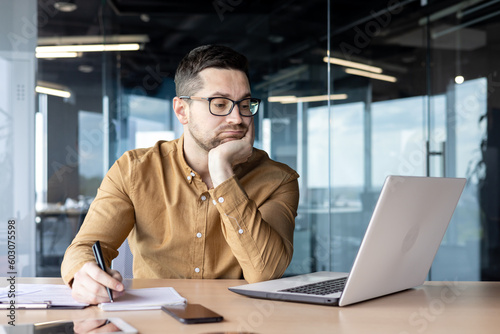 Tired and bored young man businessman, accountant, manager sitting at a table and working on a laptop and with documents. Propping his head with his hand, he looks disappointedly at the monitor