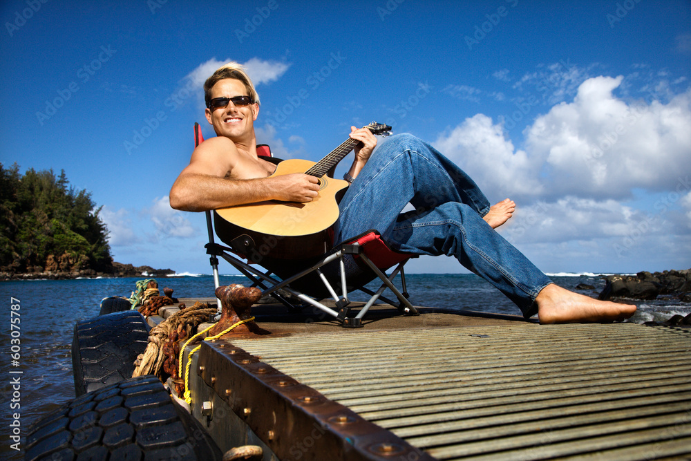Shirtless young man wearing sunglasses, sitting on a pier and playing a guitar and smiling. Horizontal shot.