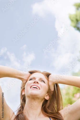 Image of a young woman outdoors with hands on her head, looking up and away from the camera. Head and shoulders viewable. Vertically framed shot.