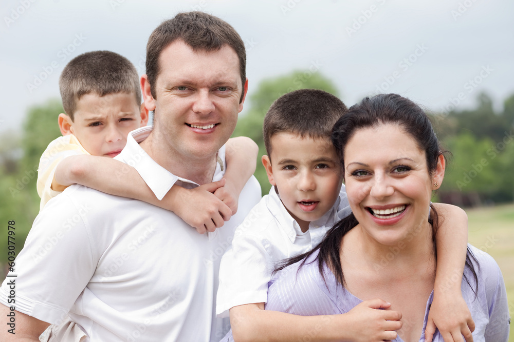 Beautiful couple carrying their kids on their back and looking at camera on natural background Beautiful couple carrying their kids on their back and looking at camera on natural background