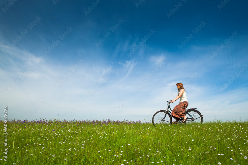 Happy young woman on a green meadow riding a bicycle - obrazy, fototapety, plakaty 