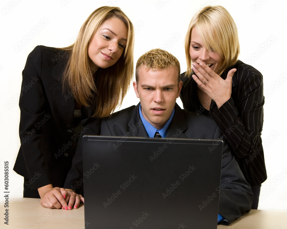 One business man sitting at desk in front of a computer with two woman coworkers standing behind him all looking at computer screen One business man sitting at desk in front of a computer with two woman coworkers standing behind him all looking at computer screen