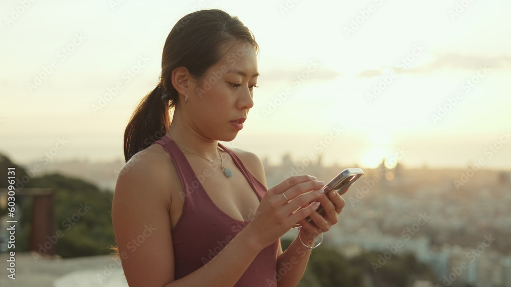 Clopse up, happy athletic woman using mobile phone while standing at lookout point at dawn