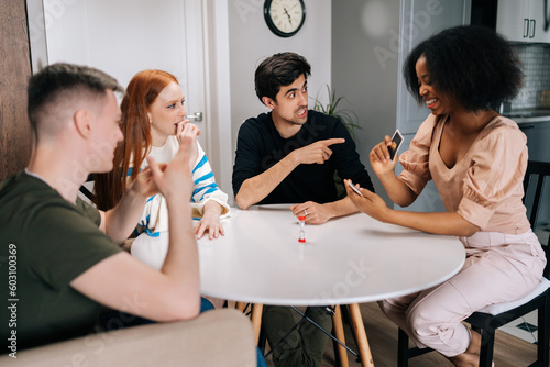 Cheerful multiethnic friends having play cards games for time enjoying board games activity for entertainment at home. Cheerful diverse men and women playing UNO board game sitting at table.