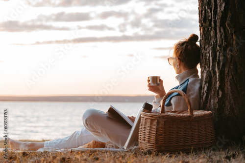 Woman drink tea or coffee from thermos read book having picnic at lake photo