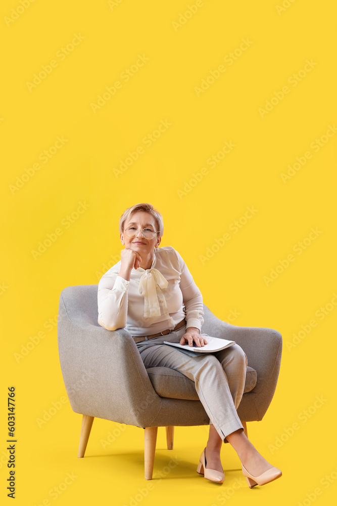 Mature psychologist with notebook sitting in armchair on yellow background Mature psychologist with notebook sitting in armchair on yellow background