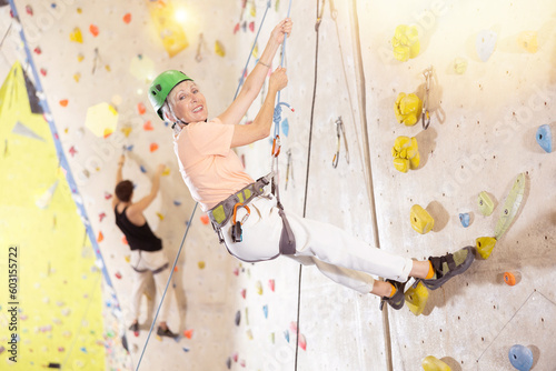 senior female climber in helmet and with safety rope hangs near climbing wall in gym. dynamic sports