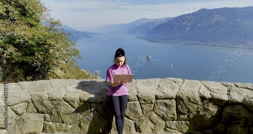 Working Outdoors with a Notebook a Woman in Fitness Clothes is Standing at the Vista Point and Drone is Revealing a Beautiful Panorama of River and Forest in Oregon State at Summer