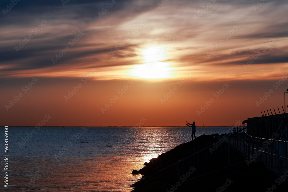 A figure casting a fishing rod out to sea whilst standing on rocks at sunset at Brighton in Melbourne
