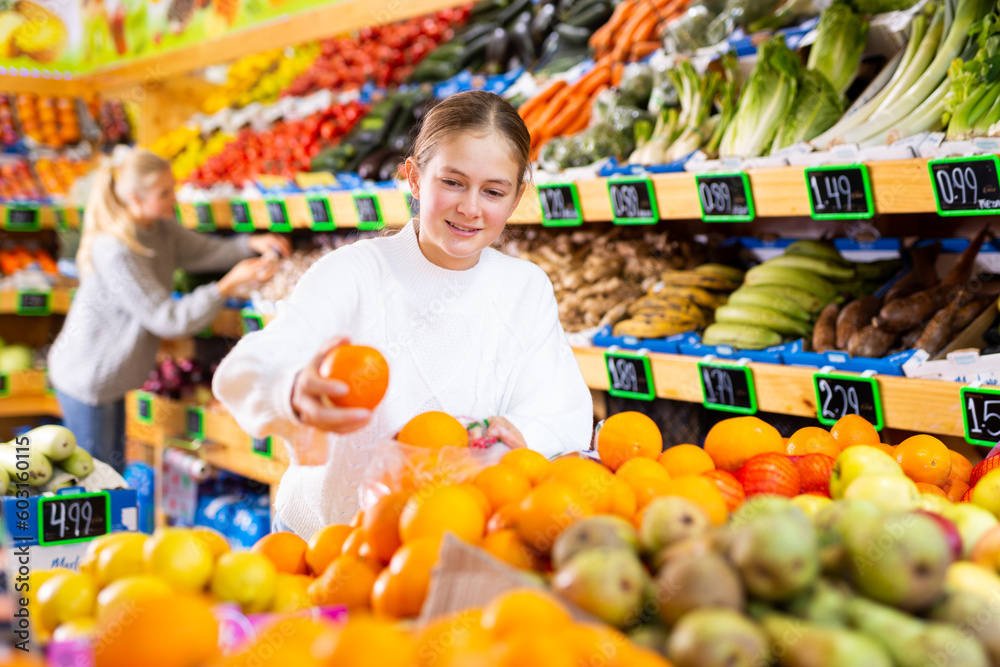 Cheerful teen girl shopping for organic fruits, choosing sweet ripe oranges in local food store ..