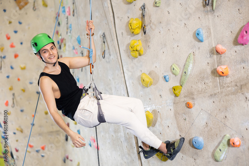 Sporty cheerful young male climber in helmet using safety belts and helmet going down after training in bouldering gym