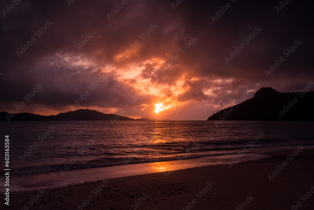 The sun showing through the clouds with the ocean waves breaking onto the shore on a tropical island