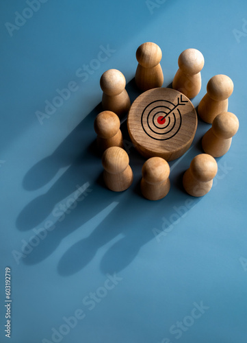 Фотография Target icon on round wood block surrounded with small wooden human figures made a crown shape shadow on blue background