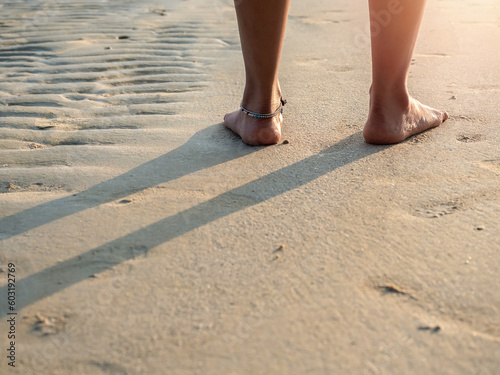 Back side of barefoot woman in white pants with folded legs standing on clean sandy beach in the warm sunlight at sunset with copy space. Summer travel, leisure destination, and vacation concepts.