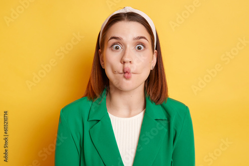 Funny optimistic woman wearing green jacket making fish lips grimacing having fun looking at camera with big eyes posing isolated over yellow background.