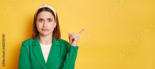 Confused puzzled woman wearing green jacket posing isolated over yellow background pointing away at copy space, advertisement area mockup. photo