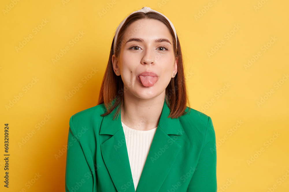 Positive cute woman wearing green jacket posing isolated over yellow background showing tongue out having fun disobedient behavior.
