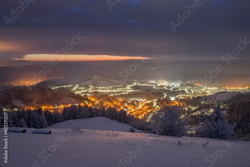Night landscape with illuminated city in the valley in winter. Banska Stiavnica, Slovakia. Church at the top of the hill. Light of houses in the valley