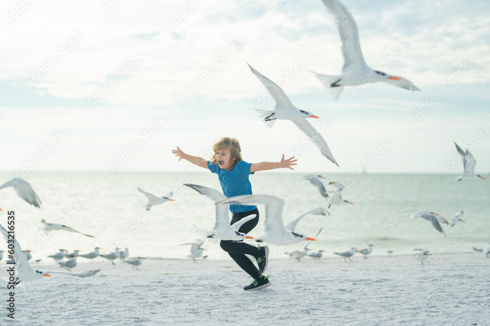 Naklejka premium Excited kid running on beach. Child run toward group of birds on sea beach. Kid running to the birds. Happy cute child running near ocean on warm sunny day.