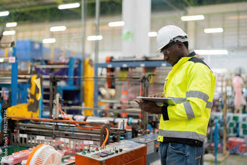 African engineer inspecting work in industrial business Wear uniforms and helmets. Background with beautiful bokeh. Holding a tablet. There is a machine to control the production of steel and plastic.