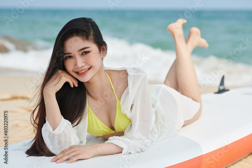 Joyful smiling young woman lying on paddleboard, beach activity concept
