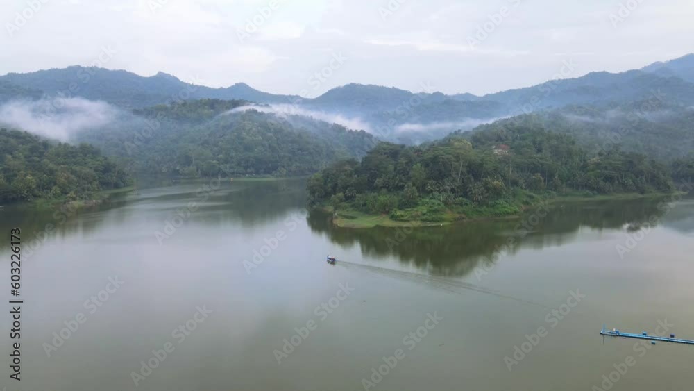 Aerial view of tranquil lake with misty forest in the morning with a boat crossing on it. Boat leave wave trails on the water surface. Reflection of the sky in the water - Shot with a rotation motion. Aerial view of tranquil lake with misty forest in the morning with a boat crossing on it. Boat leave wave trails on the water surface. Reflection of the sky in the water - Shot with a rotation motion.