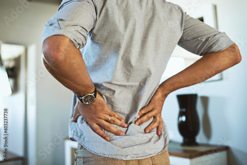 Hands, back pain and injury with a man standing in the living room of his home, holding his spine in discomfort. Health, medical and anatomy with a male person in a house feeling a muscle cramp