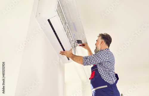 AC installation and maintenance service worker repairing or installing new air conditioner at home. Senior man in uniform workwear doing something with screwdriver in white air conditioner on wall