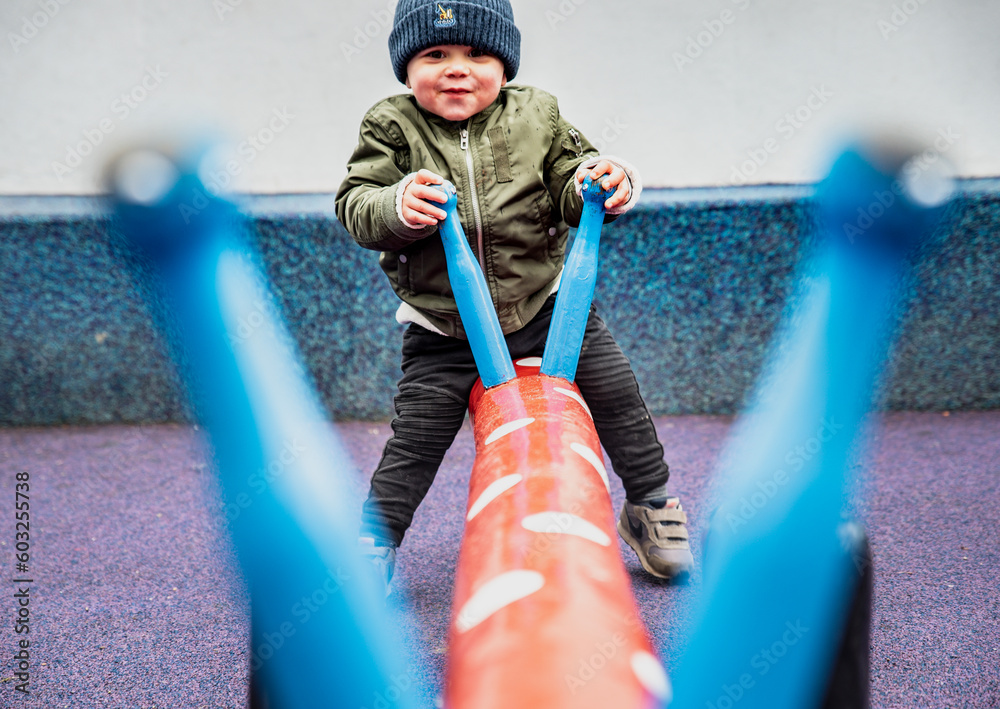 Happy child seen through the handles playing on a seesaw in a colourful playground. Playful kid outdoor on a dandle board conveys the importance of physical activity, recreation time and mental health - obrazy, fototapety, plakaty 