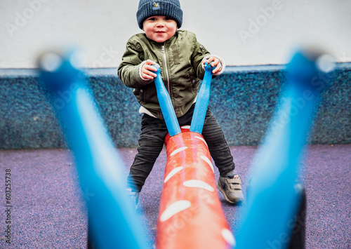 Happy child seen through the handles playing on a seesaw in a colourful playground. Playful kid outdoor on a dandle board conveys the importance of physical activity, recreation time and mental health photo
