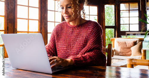 One middle age woman using computer at home with serene expression and red sweater. Wooden chalet interior house. Table workplace alternative office job business. People surfing the net alone. Travel photo