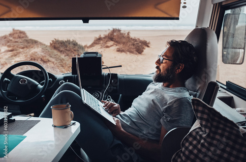 Traveler enjoy relax time in indoor leisure activity working and using laptop sitting on passenger seat inside a camper van motorhome parking in front of a beach. Digital nomad tourist lifestyle