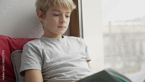 Teenager sitting next to window alone and is reading a book at home. Boy is looking out of the window. Emotional concept
