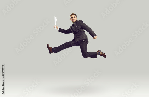 Overjoyed young Caucasian man entrepreneur in pose of running person smiles at moment of jump, rejoicing at conclusion of business contract or offer from employer located in gray studio