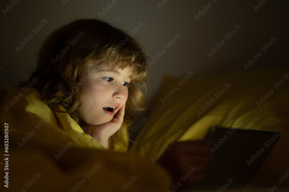 Kid lying in bed at night and playing digital tablet. Child watching tablet. Child in bed at night watching tablet, face illuminated by light of screen. Kid with tablet on bed watching cartoons. Kid lying in bed at night and playing digital tablet. Child watching tablet. Child in bed at night watching tablet, face illuminated by light of screen. Kid with tablet on bed watching cartoons.