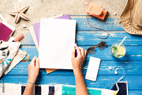 Woman hands holding blank white magazine with smart phone mockup  on beach towel