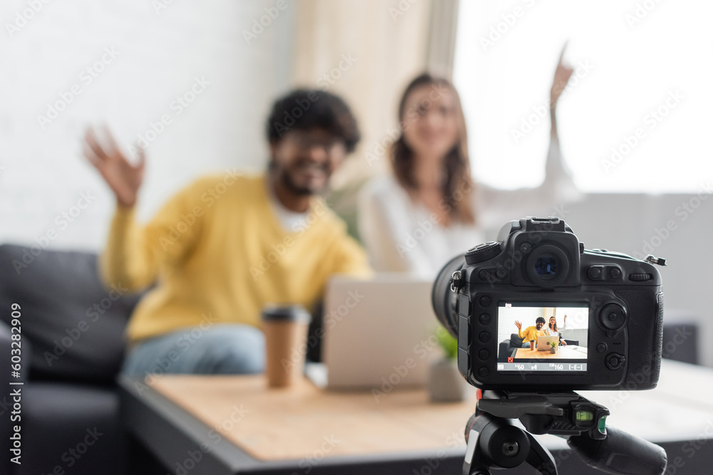 selective focus of professional digital camera near blurred interracial vloggers waving hands near laptop and coffee to go while recording podcast in radio studio