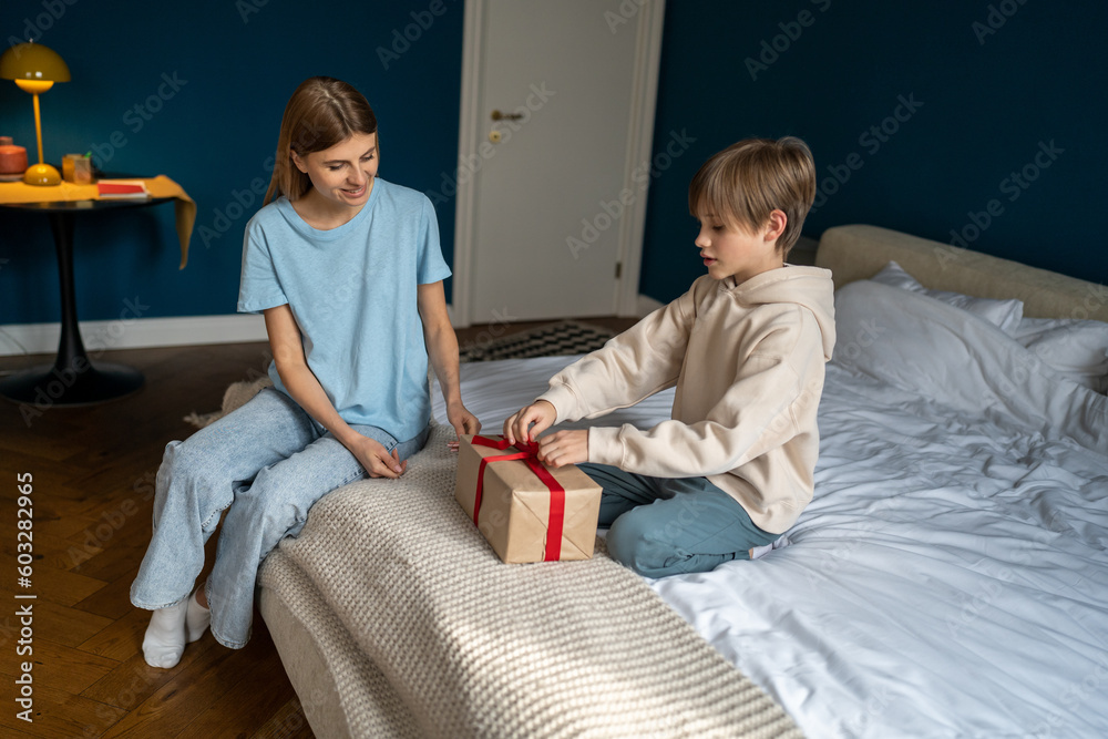 Obraz premium Cheerful kid son unwrapping gift box feeling excited getting present from caring mother, celebrating happy birthday. Loving family. Smiling mum congratulating child boy at home sits together on bed. 