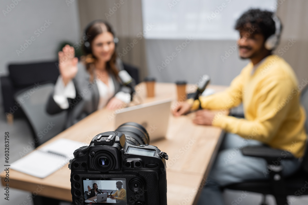 professional digital camera recording interview of woman waving hand and young indian guest sitting at table with laptop and blurred paper cups in broadcasting studio professional digital camera recording interview of woman waving hand and young indian guest sitting at table with laptop and blurred paper cups in broadcasting studio