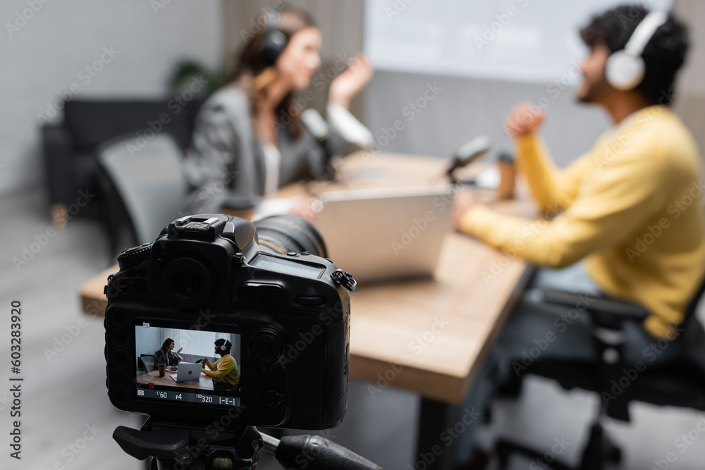 selective focus of professional digital camera recording how interviewer talking to young indian guest while sitting together near laptop and microphones on blurred background in studio