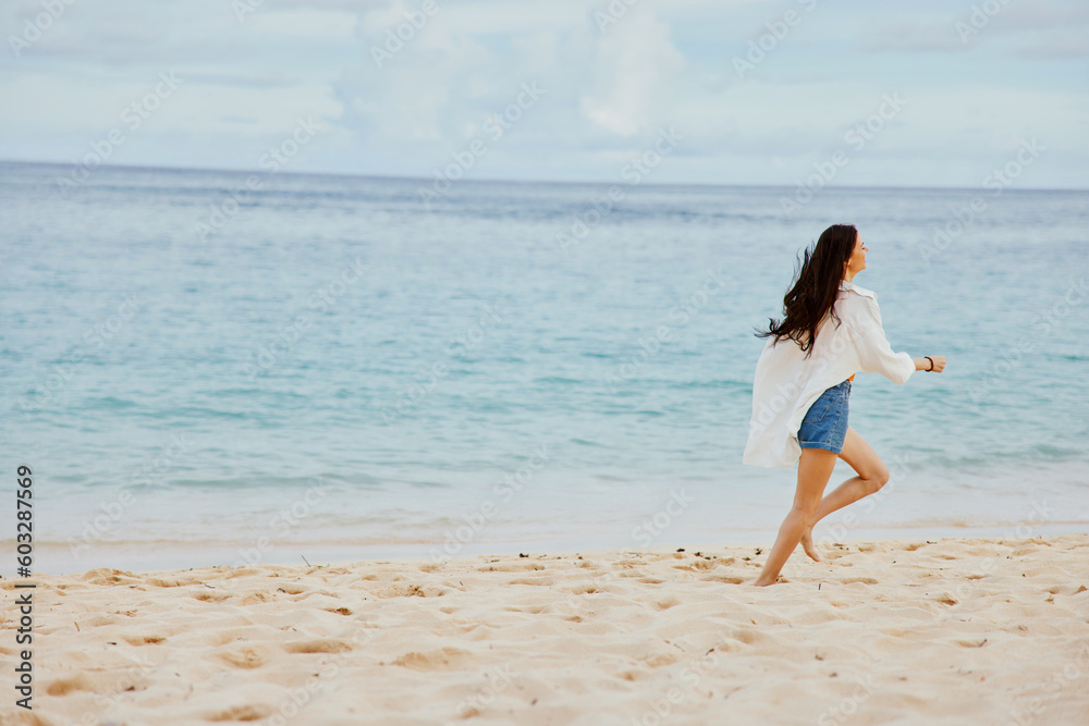Naklejka premium Sports woman runs along the beach in summer clothes on the sand in a yellow T-shirt and denim shorts white shirt flying hair ocean view, beach vacation