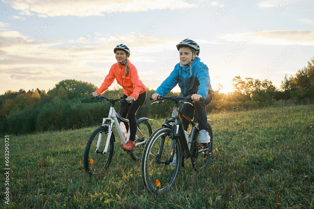 Fototapeta premium Mother and son ride bike outdoors. Happy cute boy in helmet to riding a bike in park on green meadow at sunset time. Family weekend. mothers Day 