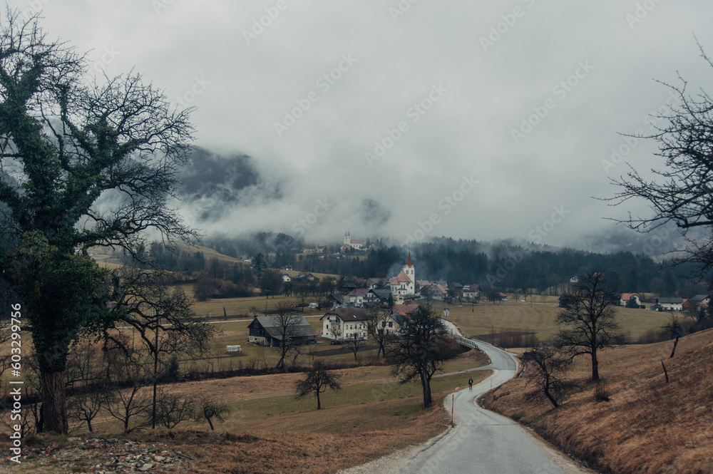 Obraz premium A captivating winter scene featuring a mountain road leading to a charming small town nestled among fog-covered mountains in Slovenia. 