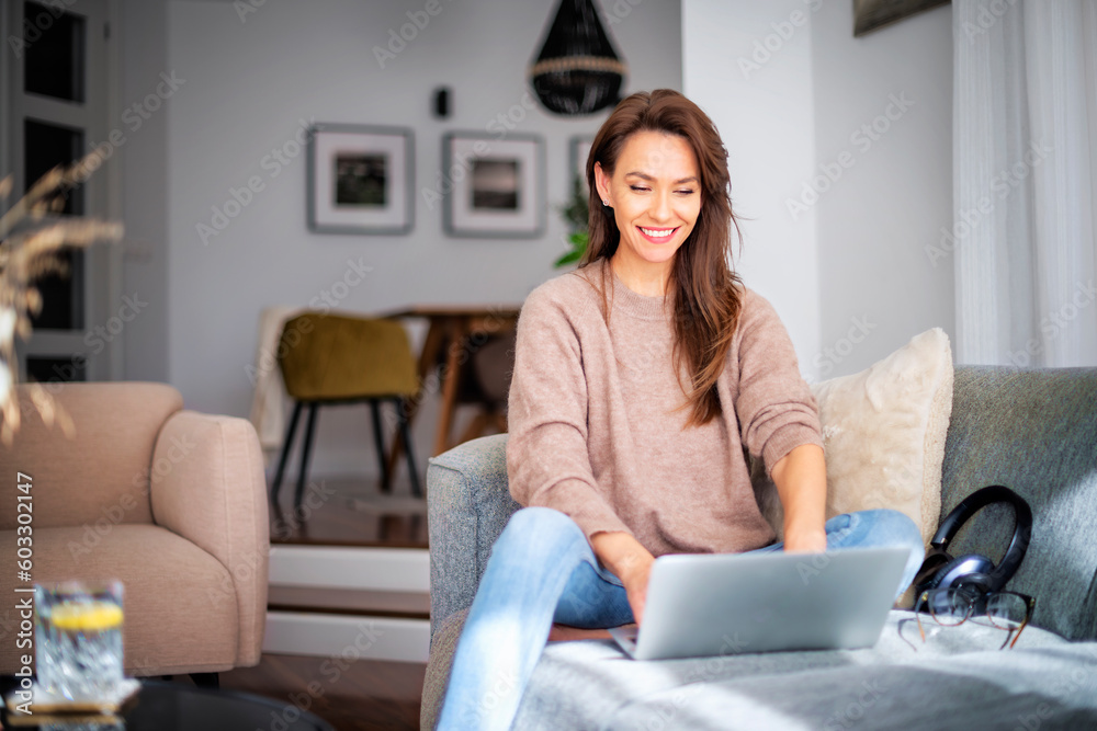Smiling caucasian woman sitting on the sofa at home and using laptop