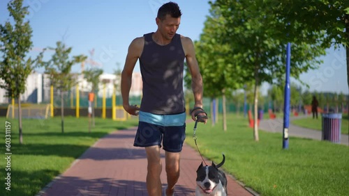 Dolly shot portrait of confident motivated sportsman jogging with dog in slow motion stopping caressing pet. Front view Caucasian man running training outdoors stroking American Staffordshire Terrier