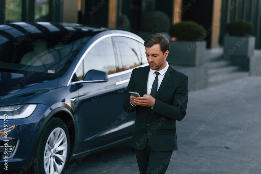 Holding smartphone. Businessman is standing near his car outdoors