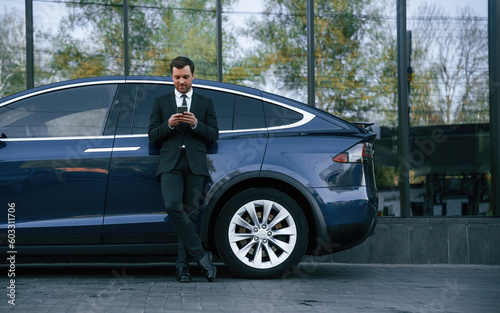 Holding smartphone. Businessman is standing near his car outdoors