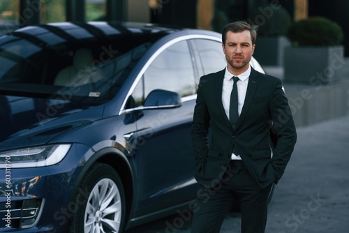 Standing, in black suit. Businessman is near his car outdoors