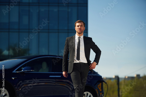 Serious facial expression. Businessman is standing near his electric car outdoors © standret